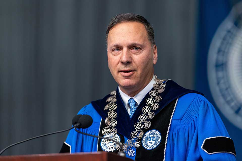 President Ron Liebowitz stands at a lectern in front of a Brandeis University banner, with others sitting behind him.