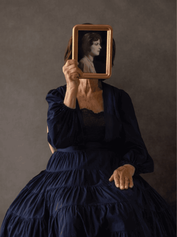 A woman wearing a flowing gown sitting and holding a photo of a black and white photo of another woman directly in front of her face.