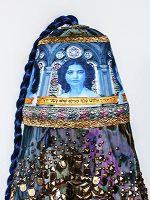 front overview of a turquoise head piece sit on a wooden pedestal. the headpiece has women's faces superimposed on it. There are golden geometric decorations attached on the bottom of the turquoise headpiece. A turquoise braid hair cascades from the top.