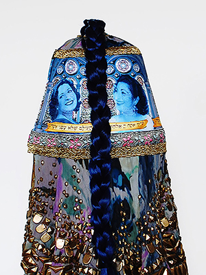 back view of a torquoise head piece sitting on a pedestal. the headpiece has women's faces superimposed on it. golden geometric decorations attached on the bottom of the turquise headpiece. Turquoise braided hair cascades from the top.