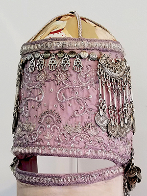 front view of a violet head piece sits on a pedestal. the head piece has two parts, they are attached on the side. the top part has embroidery decorated throughout out, silver decorations on the top, and photographic image of a girl superimposed on the top. the bottom part has silver decorations on the bottom and photographic image of a women and two children superimposed in the middle