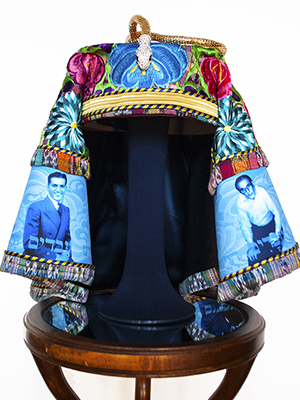 frontal view of a turquoise and red floral headpiece sits on a wooden pedestal.The fabric has photographic images of men superimposed on it. the headpiece has a golden snake at the top. 