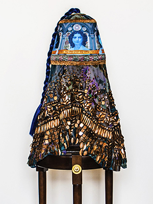 front view of a torquoise head piece sittinh on a wooden pedestal. the headpiece has women's faces superimposed on it. golden geometric decorations attached on the bottom of the turquise headpiece. Turquoise braided hair cascades from the top.
