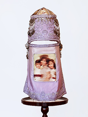front view of a violet head piece sits on a pedestal. the head piece has two parts, they are attached on the side. the top part has embroidery decorated throughout out, silver decorations on the top, and photographic image of a girl superimposed on the top. the bottom part has silver decorations on the bottom and photographic image of a women and two children superimposed in the middle