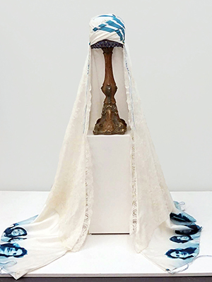 white marriage turban with blue pattern decoration on a pedestal. the long turban draped on to the floor.  photographic image of faces in blue decorated on the bottom of the marriage turban. and outside of the inner layer with the blue pattern is a white lace layer with slightly shorter length. the head part has blue stripe decorations.