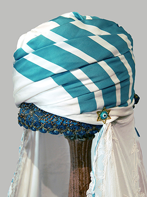 close up of white marriage turban piece with blue stripes on the fabric. A brocaded band is near the bottom of the fez cap