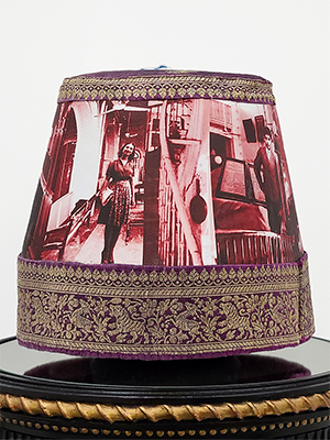 front view of a headpiece on a pedestal. photographic image of a man with background of a hall in red and white superimposed on the body of the headpiece. the top and bottom are in golden floral embroidery. on the top is a blue and white braided rope attached. 