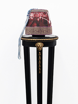 front view of a headpiece on a pedestal. photographic image of a man with background of a hall in red and white superimposed on the body of the headpiece. the top and bottom are in golden floral embroidery. on the top is a blue and white braided rope attached.