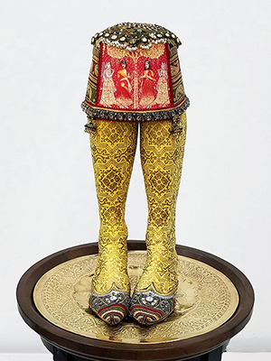 front view of sculpture on a wooden pedestal. two legs below the knees are topped with a decorated red fez cap. the legs are covered in a yellow ornate fabric, and the shoes are pointed and covered in a multicolored silky material