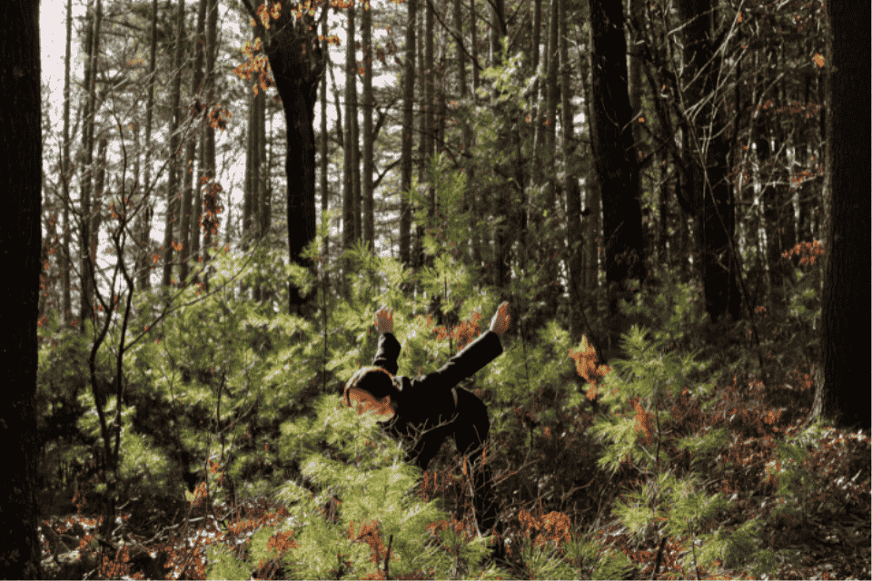 A woman in a sunny forest leaning forward with arms outstretched behind her