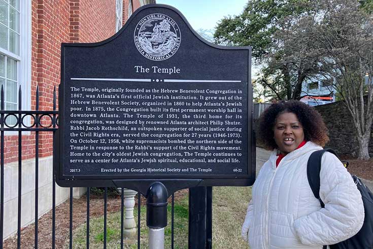 Shula Mola, wearing a white coat, is standing my a plaque outside The Temple in Atlanta.