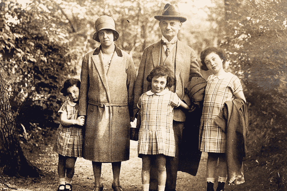 A black and white photo showing a family in the early 1900s consisting of two parents and three small children standing outdoors on a path among trees