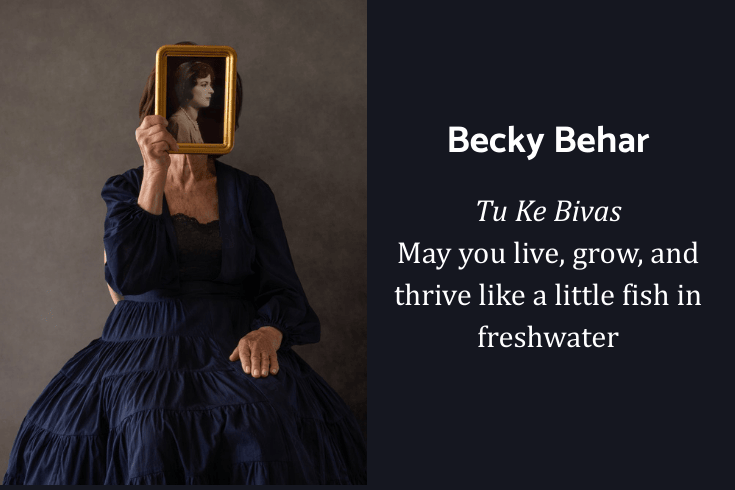 A woman wearing a flowing gown sitting and holding a photo of a black and white photo of another woman directly in front of her face. Text: Tu Ke Bivas: ‘May you live, grow, and thrive like a little fish in freshwater.’