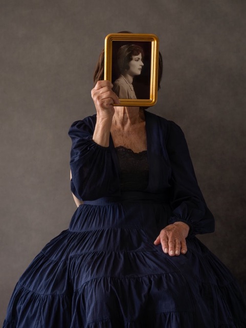 A woman wearing a flowing gown sitting and holding a photo of a black and white photo of another woman directly in front of her face