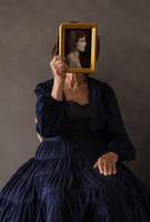A younger woman wearing a flowing gown sitting and holding a photo of a black and white photo of an older woman directly in front of her face.