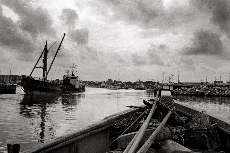 A black and white photo of a harbor from the viewpoint of sitting in a row boat with a small ship passing to the side.