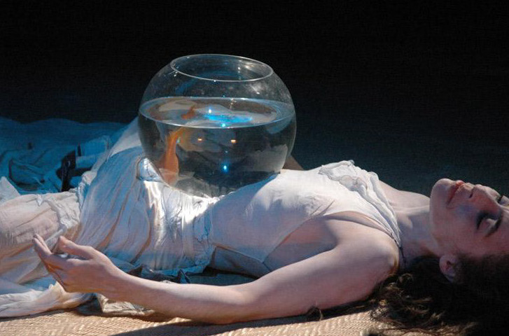 A color photograph of a woman lying on the ground on her back with a large glass, fishbowl (complete with goldfish) on her stomach.