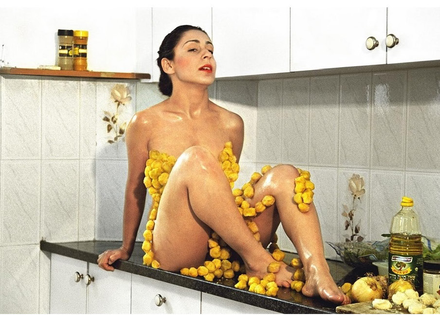A color photograph of a nude woman sitting on a kitchen counter covered in lemons. 