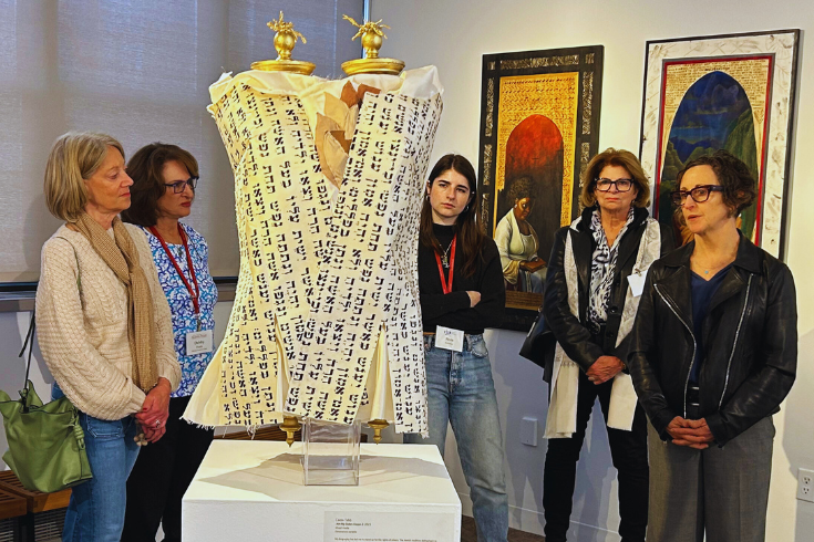 Women in an art gallery gathering about a an artwork that looks like a Torah with Hebrew writing on it. Caron Tabb, the artist, is on the right.