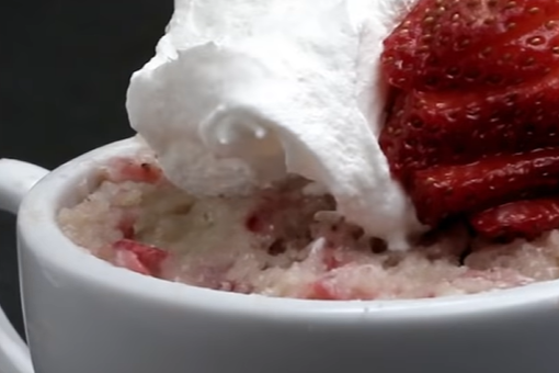 Screenshot from a video of how to make mug cakes with a close-up image of a mug cake with strawberry cake, whipped cream, and strawberries on top