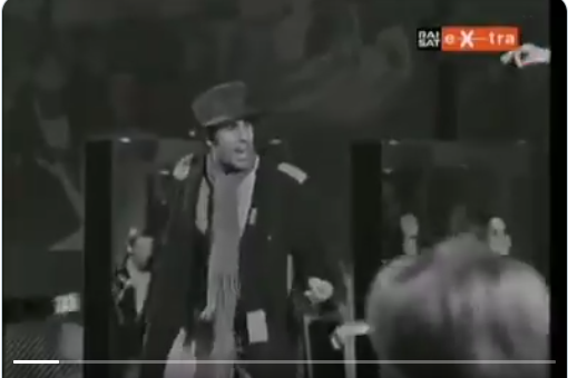 Screenshot from a black and white 70s music video with a man standing on a stage surrounded by dancers