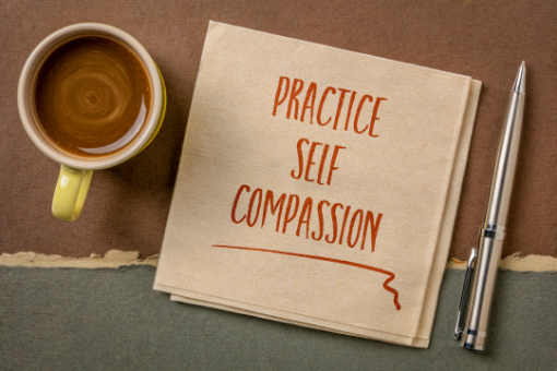 A coffee cup, a pad of paper, and a pen are on a desk. The pad of paper reads "Self-Compassion"