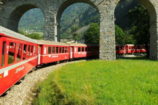Image of a red train going under an arched bridge next to grass