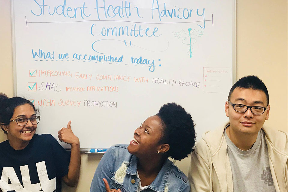 Three students sit in front of a white board that talks about the Student Health and Advisory Committee