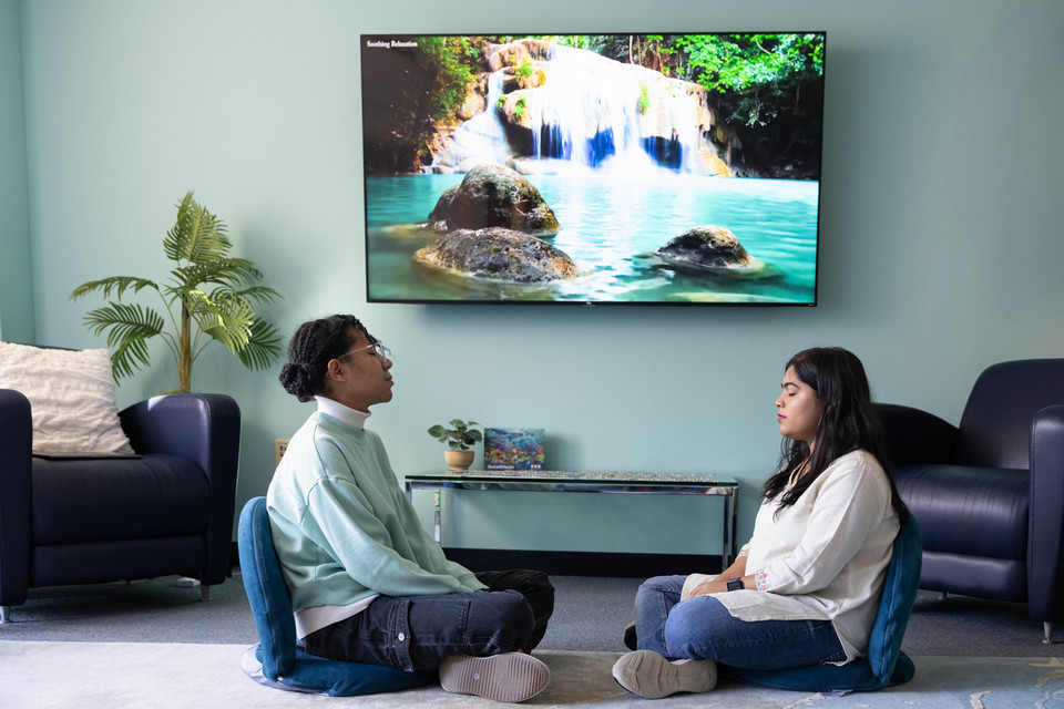 Two students meditate on the floor in front of a tv with relaxing waterscapes