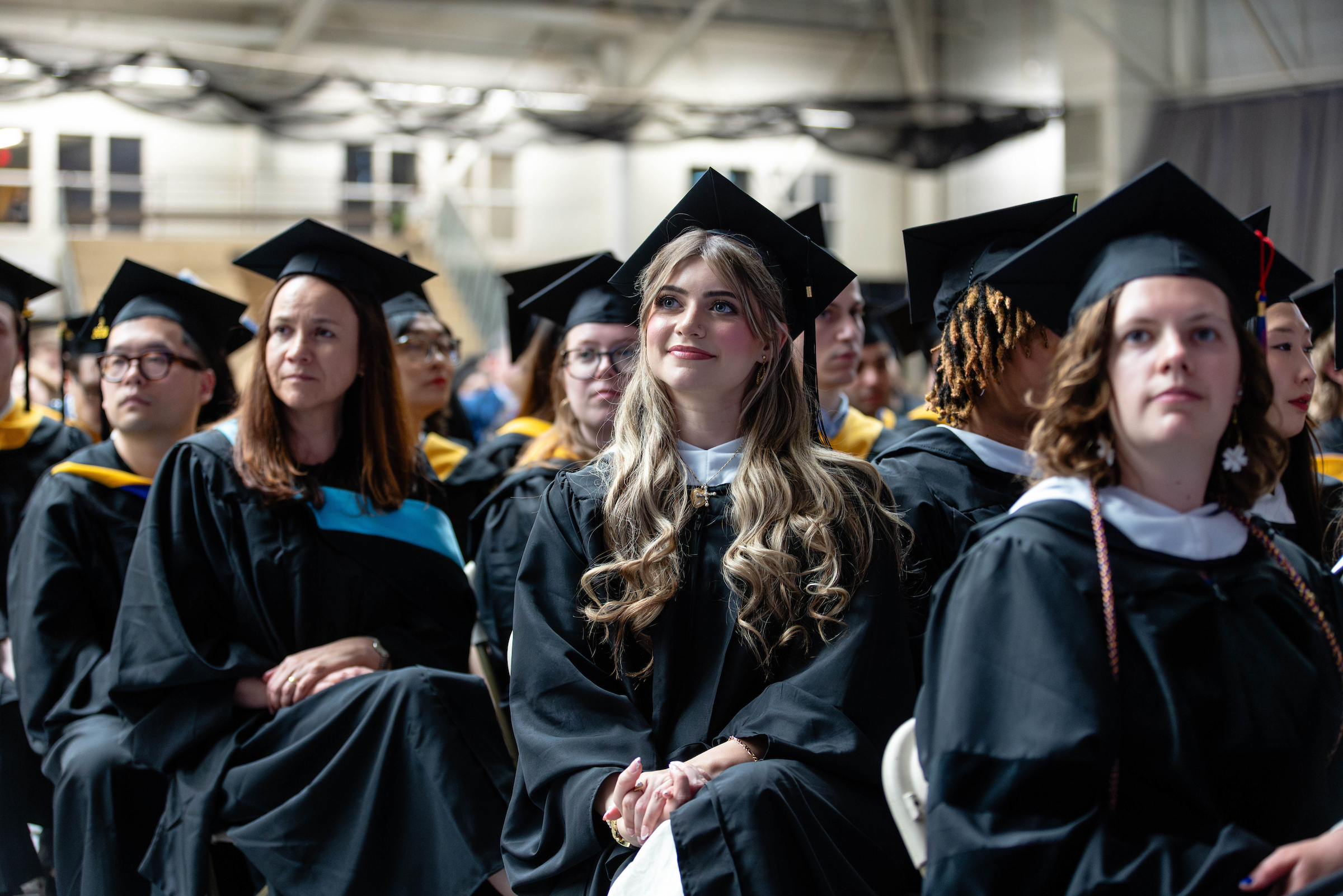 Graduates in caps and gowns seated at a 2025 commencement ceremony, with one smiling student centered in the foreground looking toward the stage.