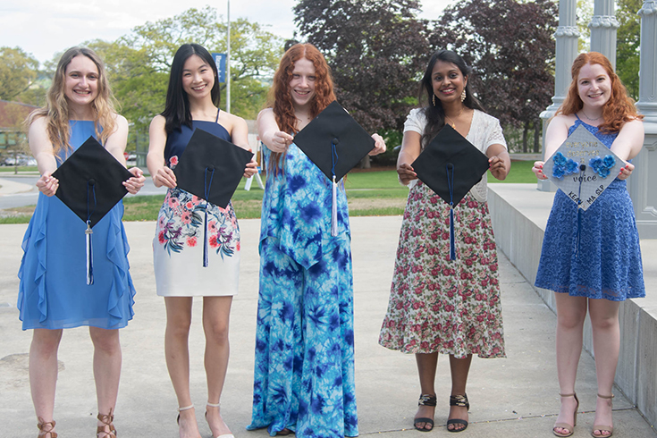 Five graduates pose for a picture holding their graduation caps out towards the camera