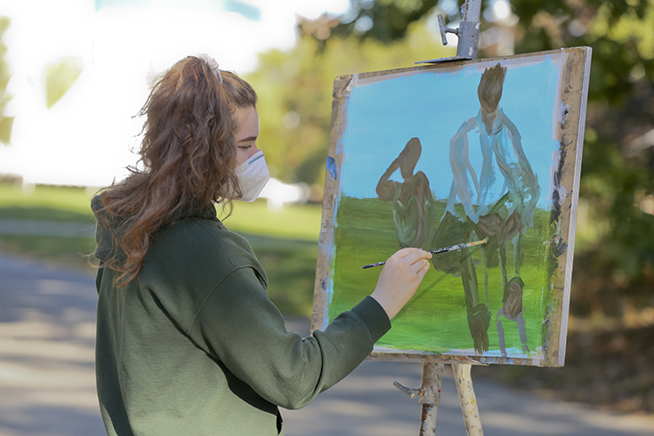 A student paints an image of two people sitting outside during an outdoor art class.