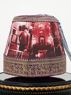 front view of a headpiece on a pedestal. photographic image of a man with background of a hall in red and white superimposed on the body of the headpiece. the top and bottom are in golden floral emboridery. on the top is a blue and white braided rope attached. 