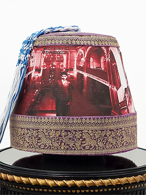 front view of a headpiece on a pedestal. photographic image of a man with background of a hall in red and white superimposed on the body of the headpiece. the top and bottom are in golden floral embroidery. on the top is a blue and white braided rope attached. 
