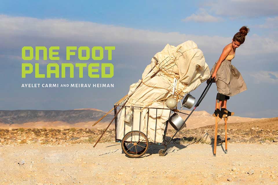 A person on stilts pulls a cart filled with fabric and pots and pans (Text: One Foot Planted, Ayelet Carmi and Merirav Heiman)