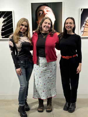 Three women standing together in an art gallery in front of a wall of photographs of women
