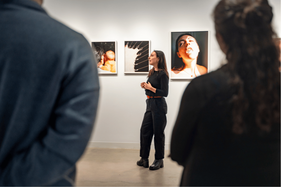 A woman standing in front of a wall of photographs of women speaking to people at an art gallery