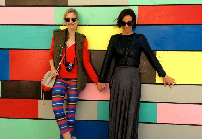 A photograph of two women standing, holding hands, and leaning against a bright, color-blocked wall.  