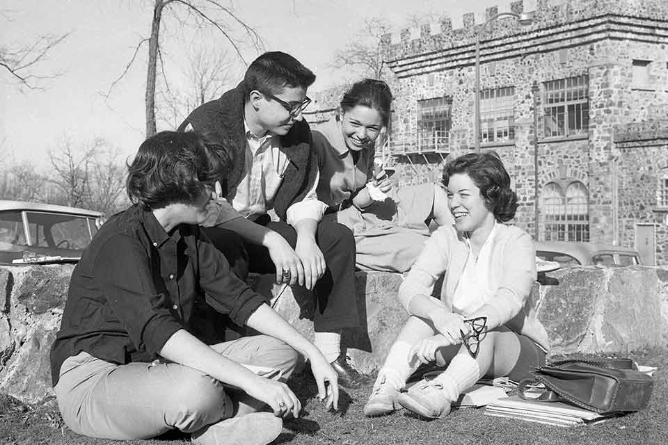 Black and white photo of four students on the Brandeis campus, the castle is seen in the background.