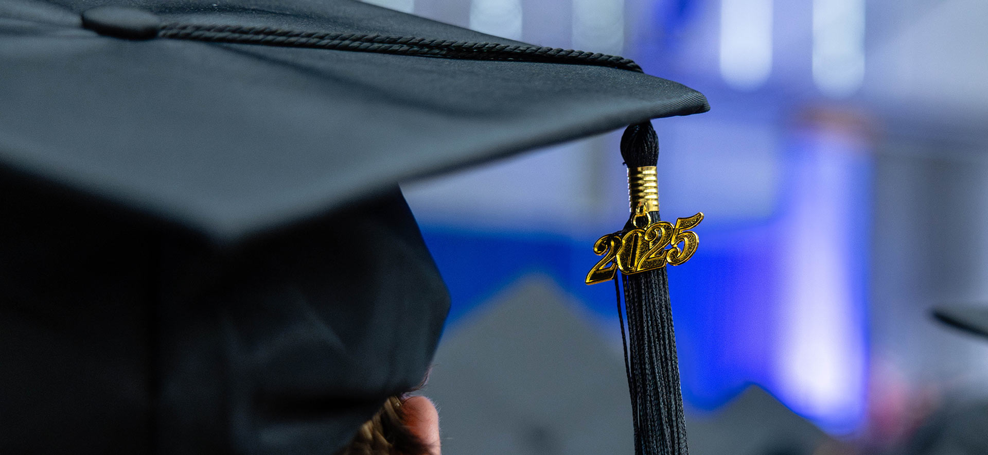 Two students dressed in caps and gowns smiling outside Gosman Sports and Convocation Center; green leaves from a tree are seen in the foreground.