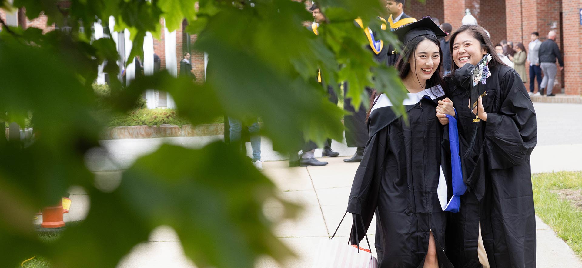 Two students dressed in caps and gowns smiling outside Gosman Sports and Convocation Center; green leaves from a tree are seen in the foreground.