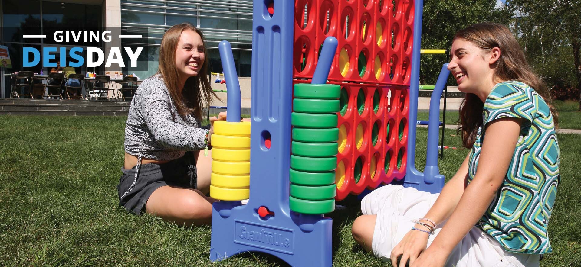 Two students sit on the ground in front of the Shapiro Campus Center playing an oversize Connect 4 game. The words "Giving DEISDAY" are in the upper left corner.
