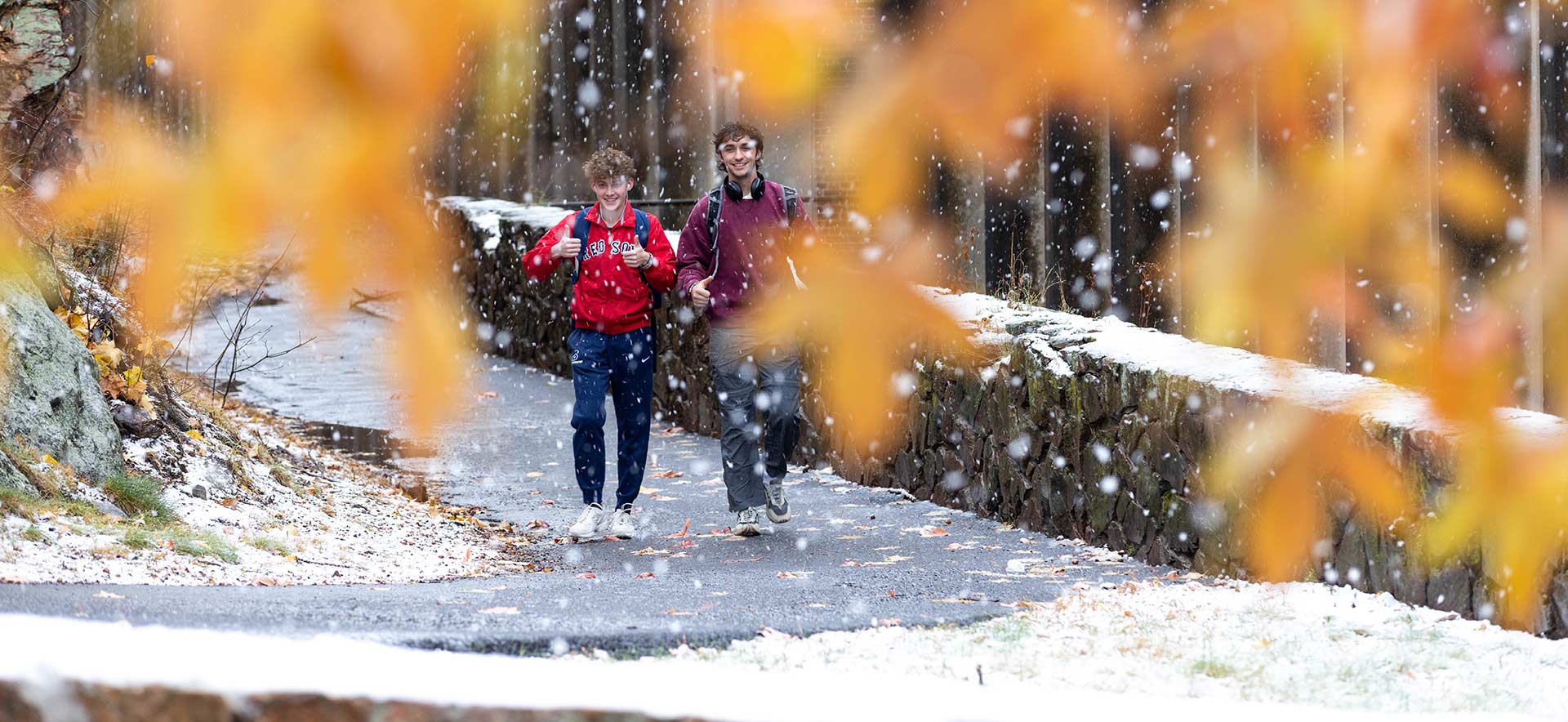 Two students walking on campus. Orange colored leaves are seen on trees and a dusting of snow is on the ground.
