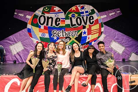 Six students, some holding flowers, pose in front of a colorful Culture X banner