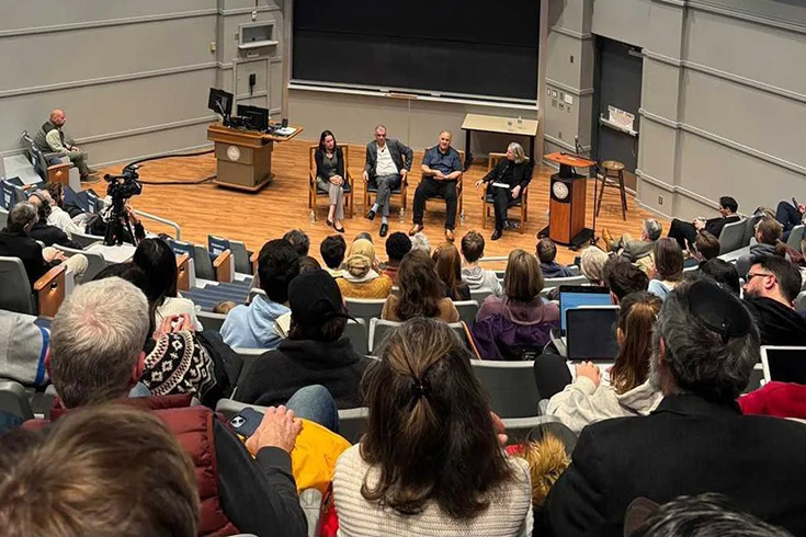 An indoor audience in stadium-style seating watches speakers seated on a stage.