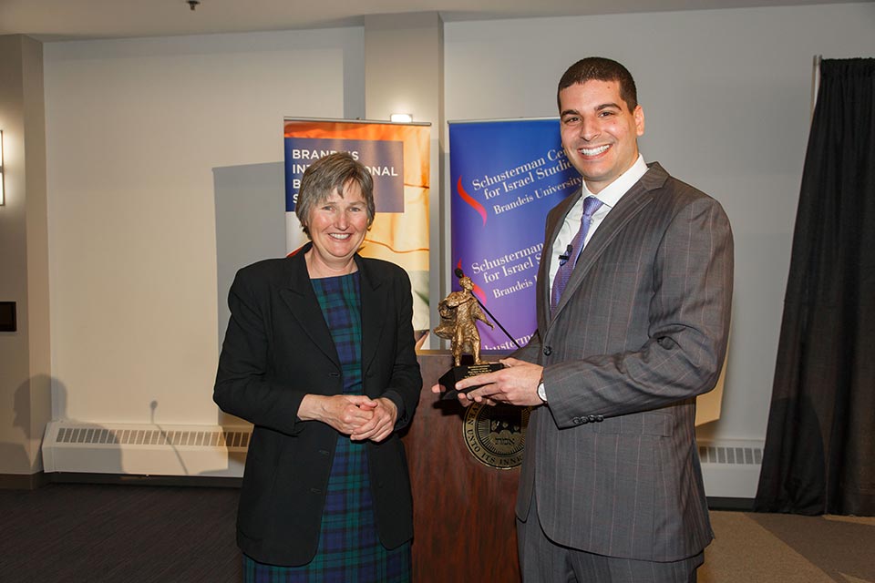 Kathryn Graddy stands next to Ohad Elhelo, who is holding the award, which is a miniature version of the sculpture of Louis Brandeis at the heart of the Brandeis University campus. Both are smiling at the camera.