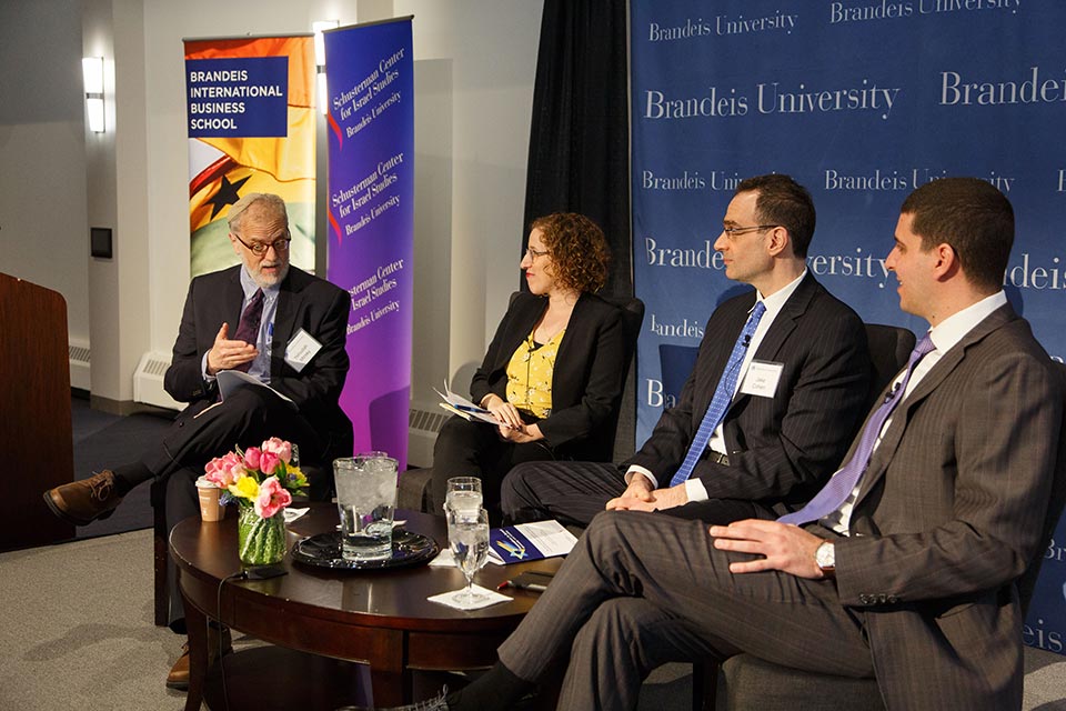 Four people sit on a stage, engaged in discussion. A man with a neatly trimmed, white beard; a red-headed woman; a tall man in glasses; and a young man with dark hair. 