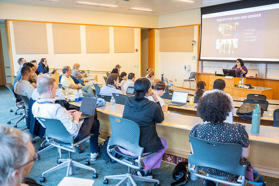 A group of people seated facing a woman at a podium with a presentation projected on a screen behind her.