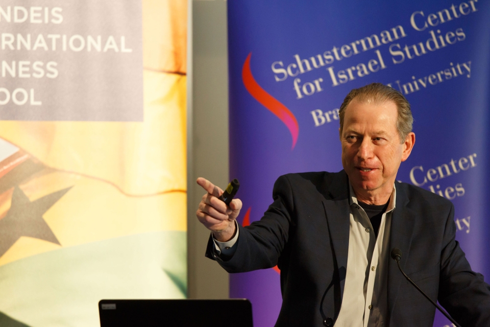 Speaker Eran Carmel at podium with arm outstretched, and purple Schusterman Center banner behind him.