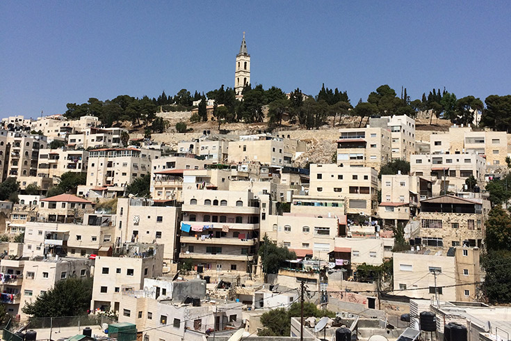 Hill in Jerusalem stacked with small, stone homes, capped by a tower stretching into a blue sky above.